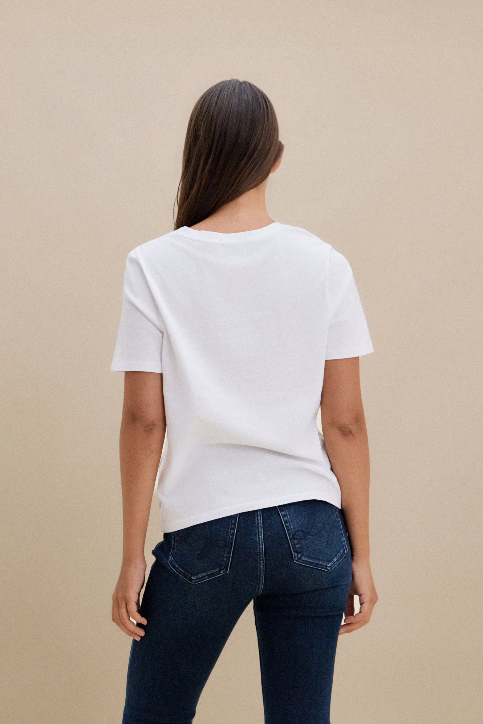 Woman wearing a plain white t-shirt and blue jeans against a beige background 