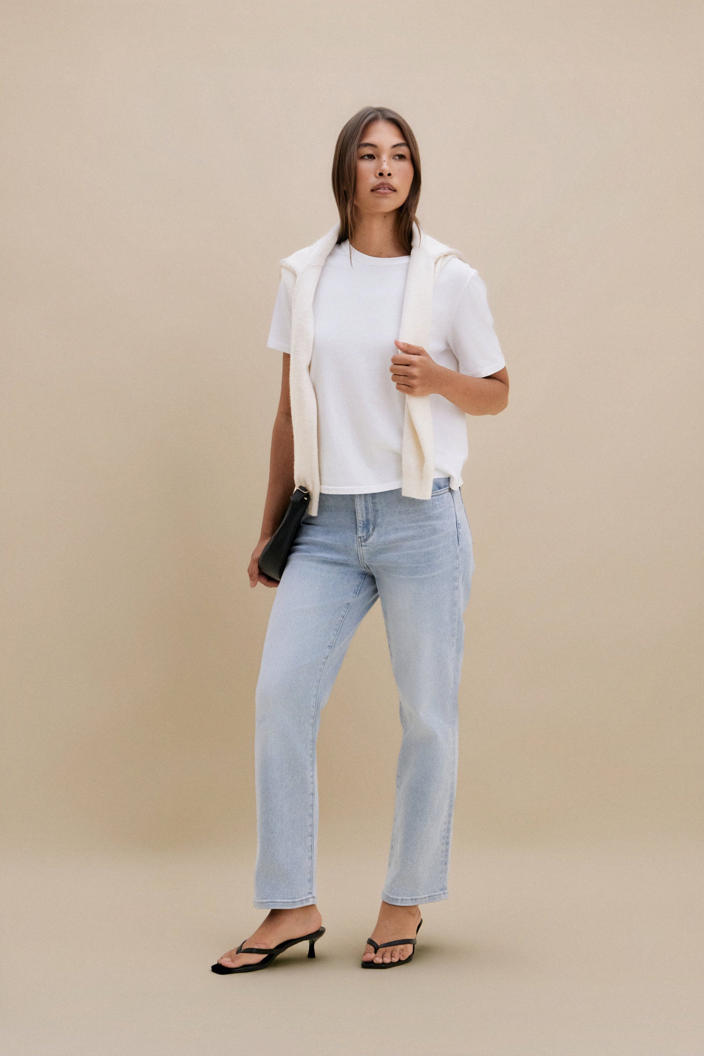 Woman wearing a white shirt, beige vest, and light blue jeans against a beige background 