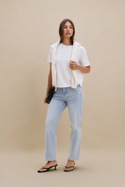 Woman wearing a white shirt, beige vest, and light blue jeans against a beige background 