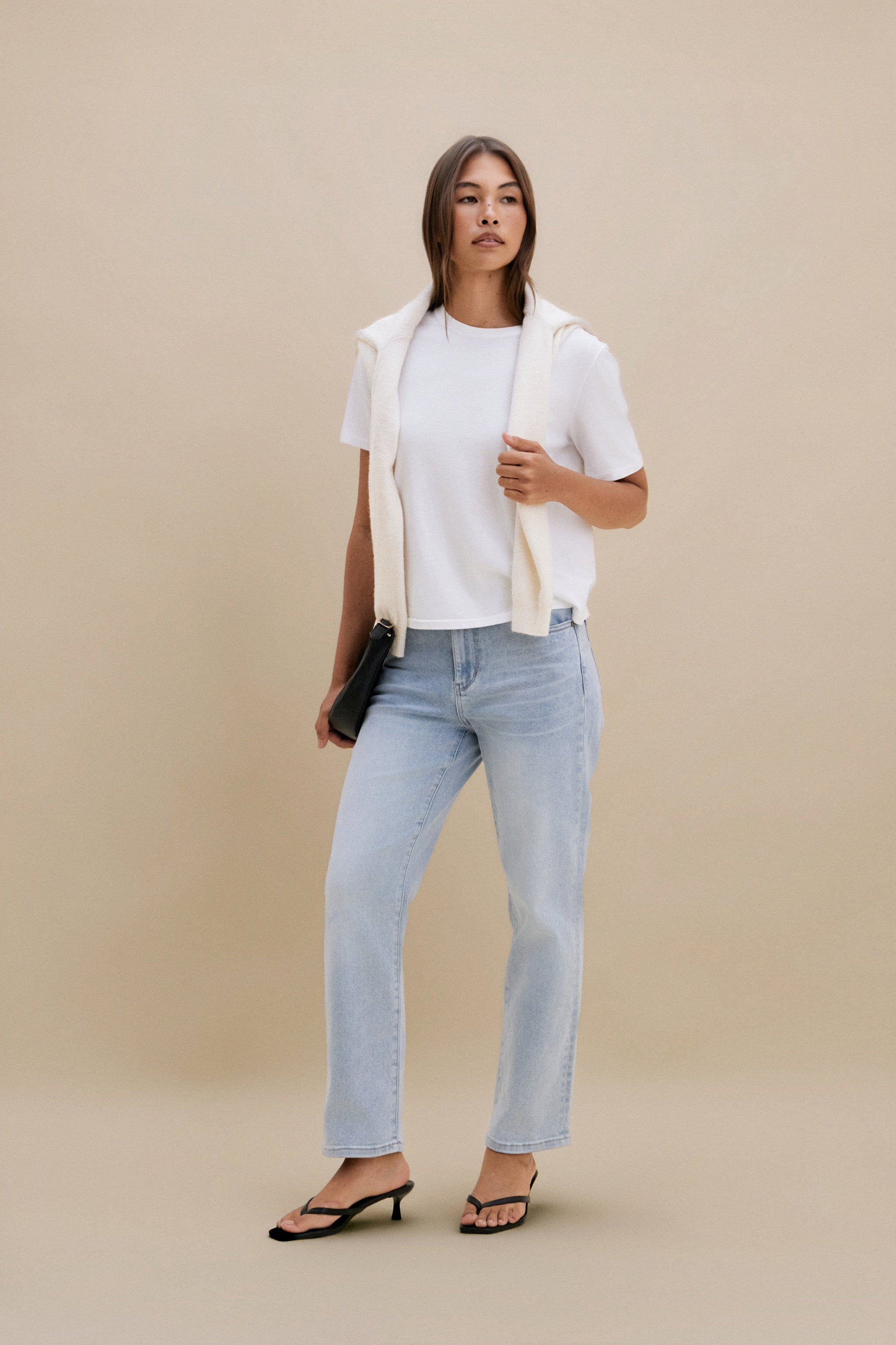 Woman wearing a white shirt, beige vest, and light blue jeans against a beige background 