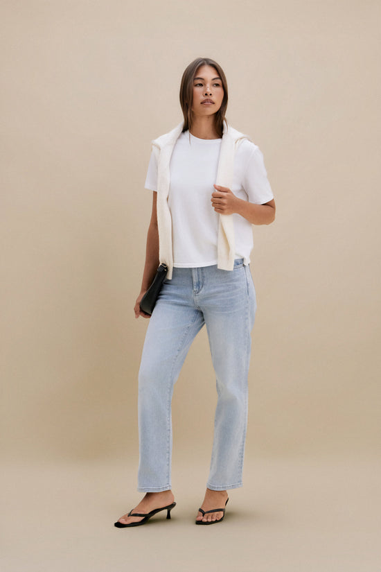 Woman wearing a white shirt, beige vest, and light blue jeans against a beige background 