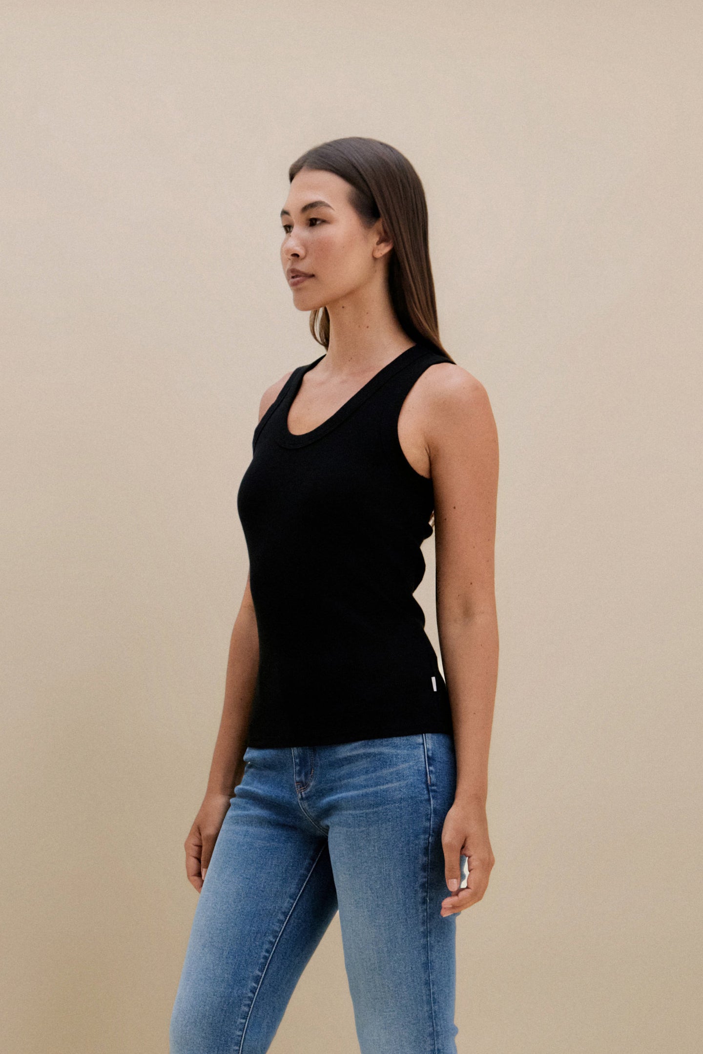 Woman wearing a black tank top and blue jeans against a beige background