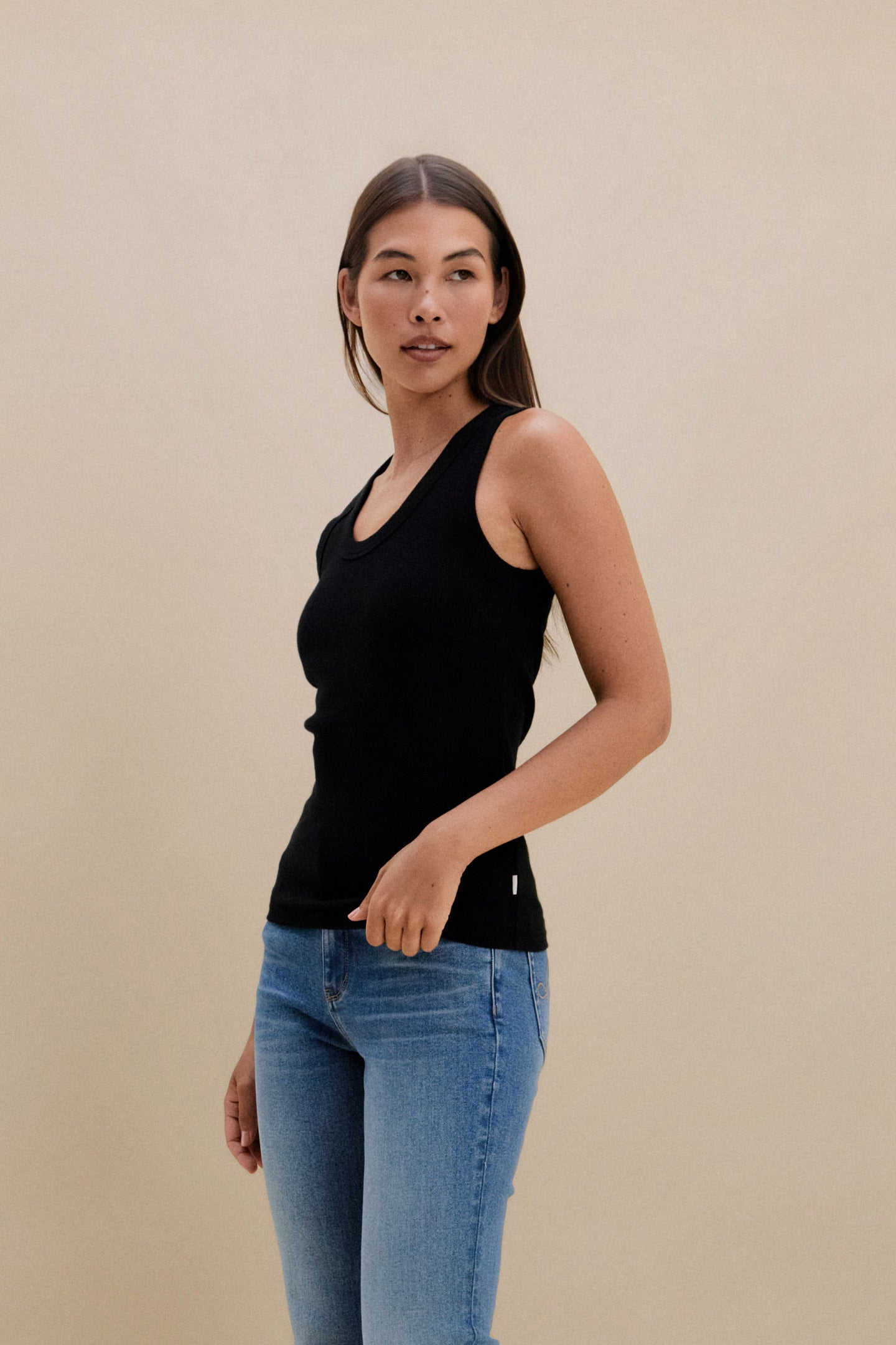 Woman wearing a black tank top and blue jeans against a beige background | model-size: 4