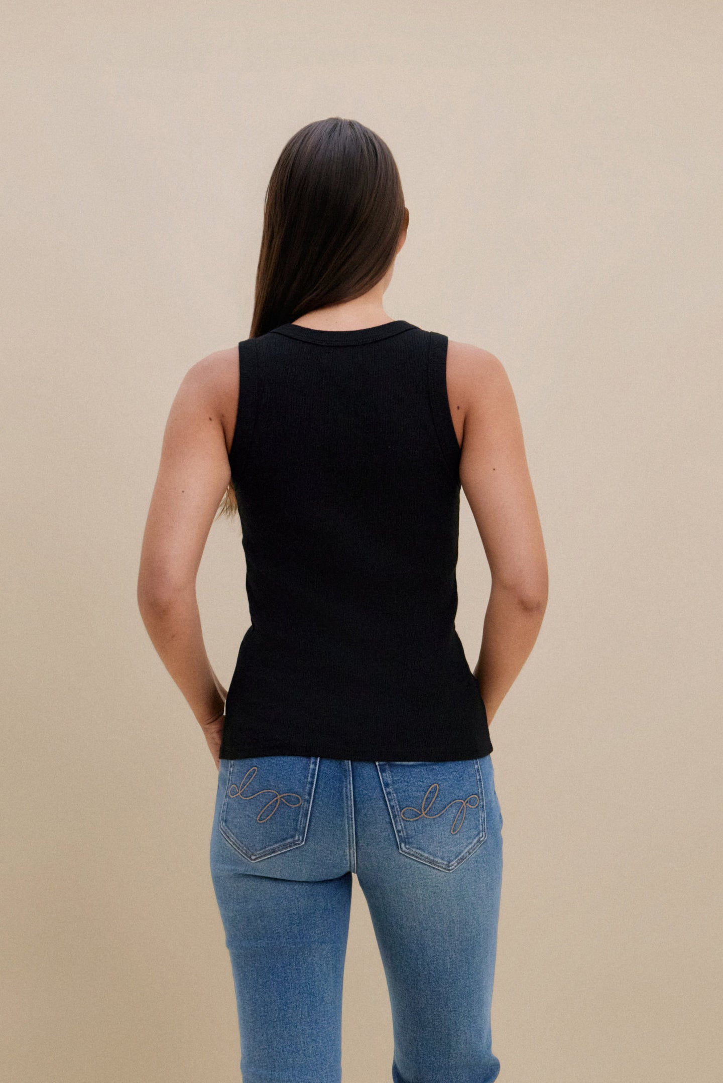 Person wearing a black sleeveless top and blue jeans against a beige background