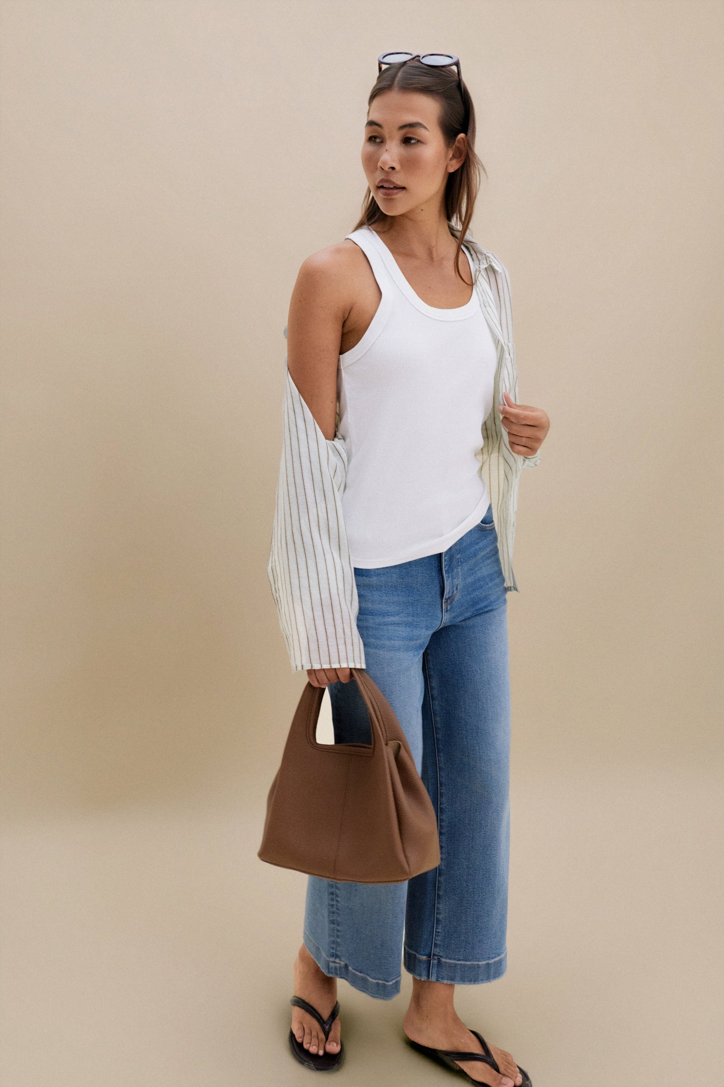 Woman wearing a white tank top, striped cardigan, and blue jeans, holding a brown handbag against a beige background.