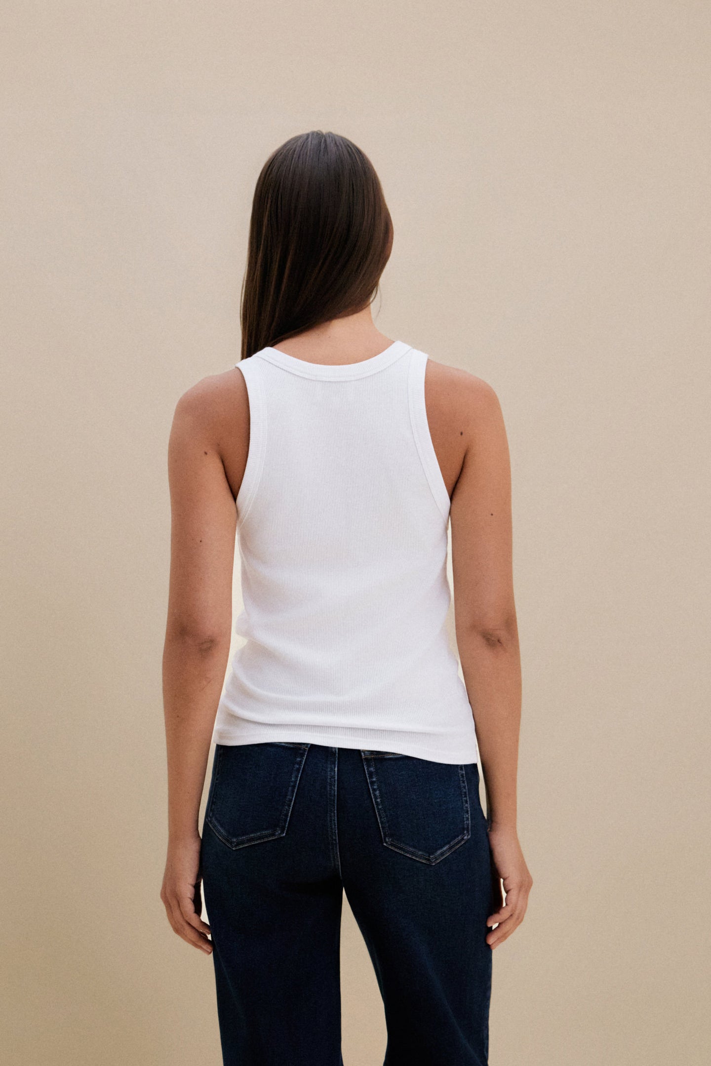 Person wearing a white tank top and dark jeans against a beige background