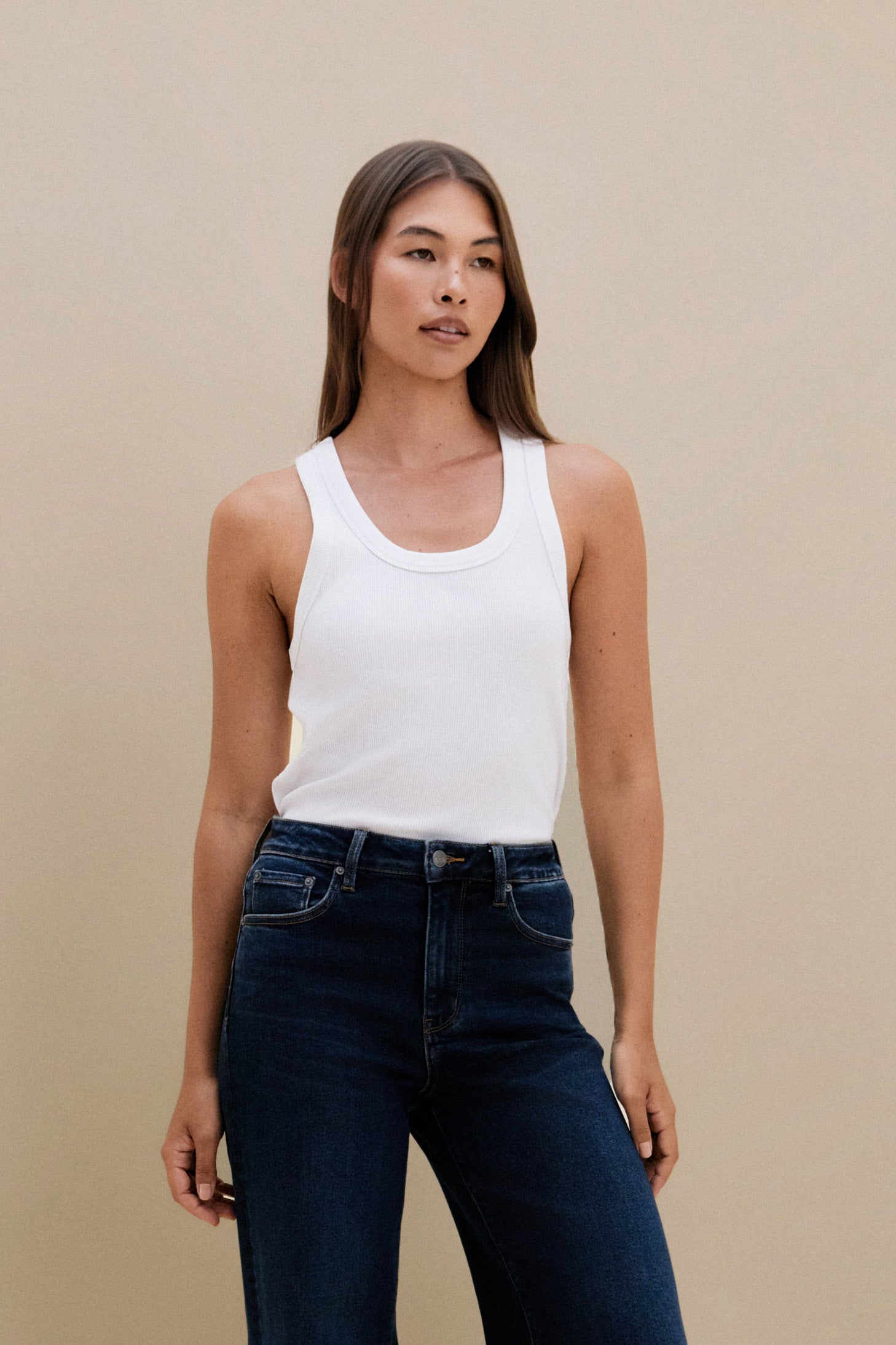 Woman wearing a white tank top and dark jeans against a beige background