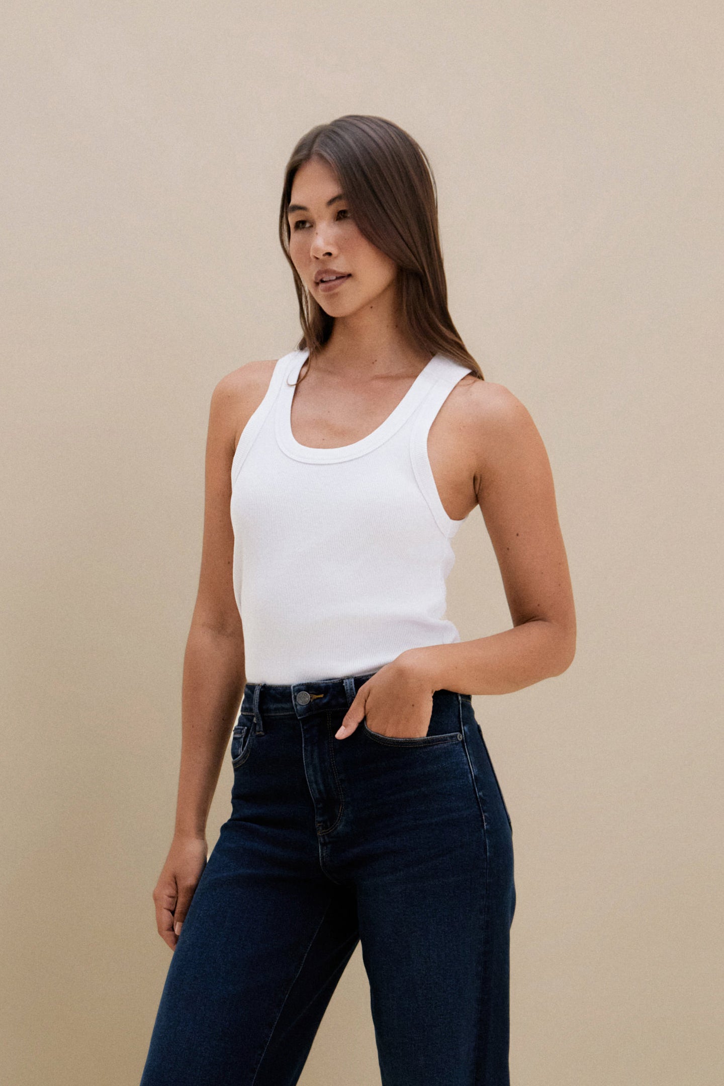Woman wearing a white tank top and dark jeans against a beige background
