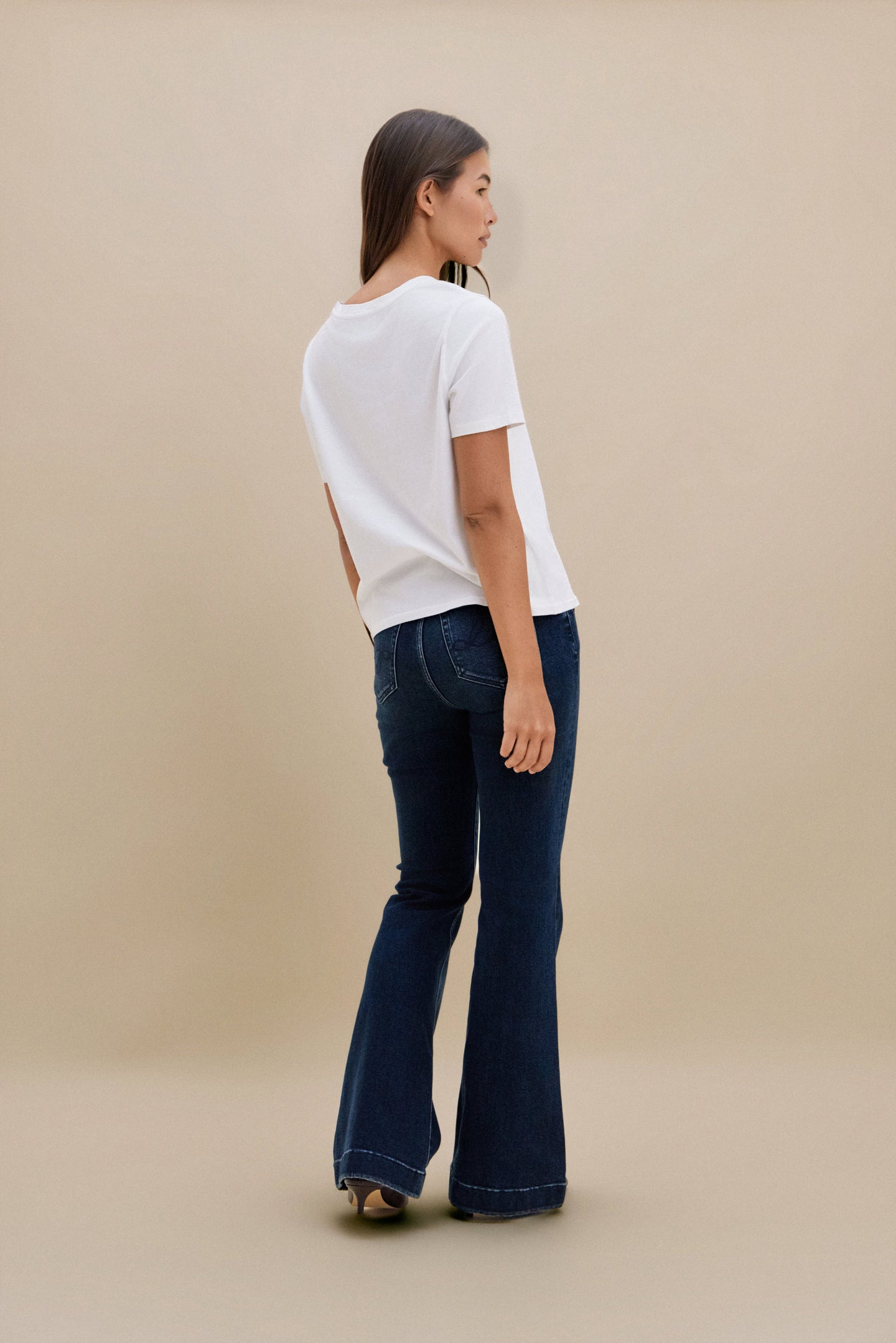 Woman wearing a white top and dark blue flared jeans against a beige background