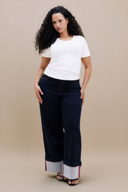 A woman modeling dark wide-leg jeans with rolled cuffs showing a light inner lining, paired with a white t-shirt, against a solid beige studio background.