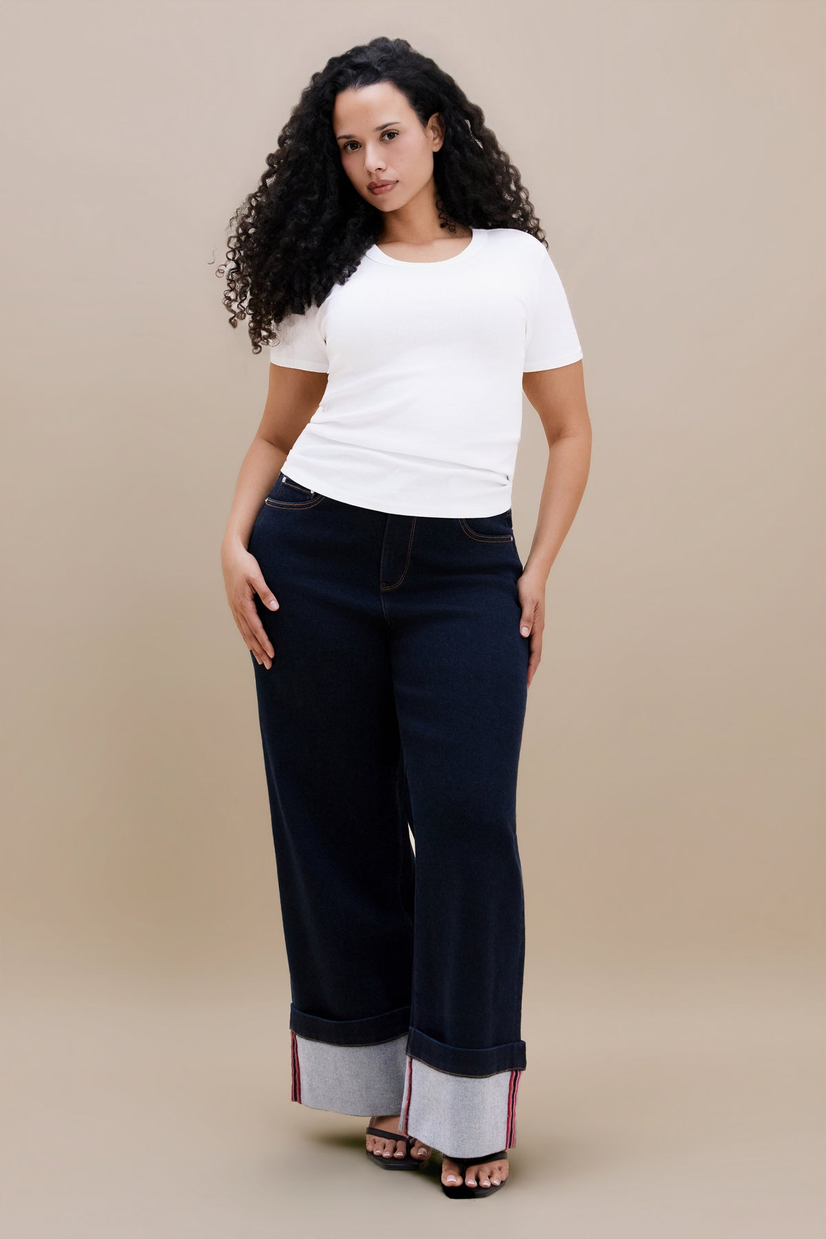 A woman modeling dark wide-leg jeans with rolled cuffs showing a light inner lining, paired with a white t-shirt, against a solid beige studio background.
