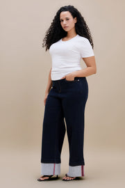 A woman modeling dark navy wide-leg jeans with light gray cuffed hems and thin red and blue stripe, paired with a white t-shirt and black sandals, against a solid beige background.