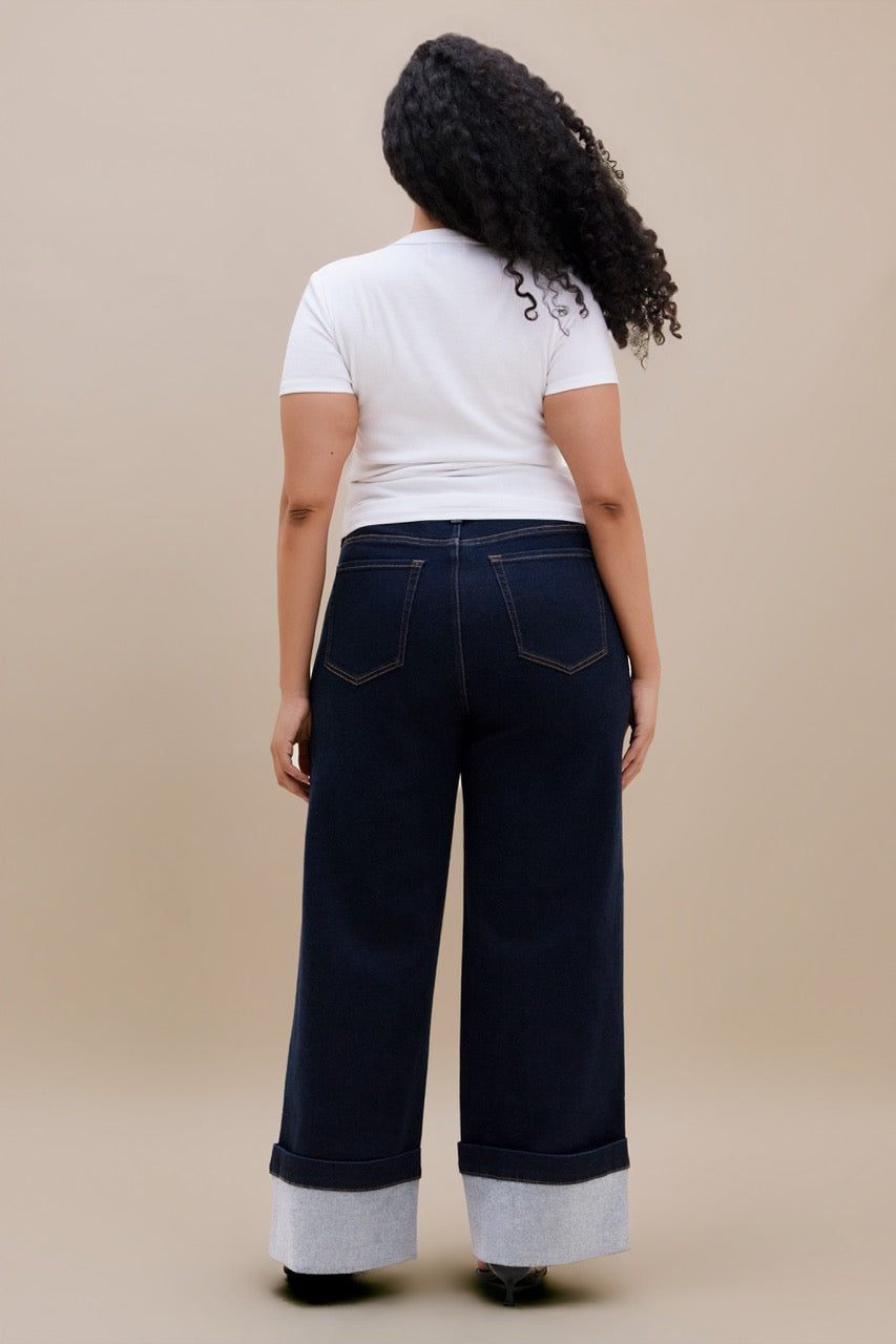 A woman viewed from behind wearing a white short-sleeve t-shirt and dark navy wide-leg cuffed jeans against a solid beige studio background.
