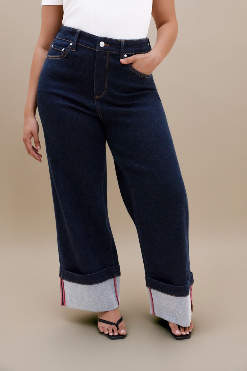 A model wearing dark navy blue wide-leg cropped jeans with rolled cuffs showing a light gray lining, styled with a white top and black sandals against a solid beige background.