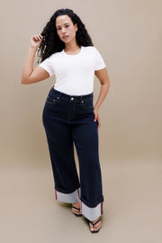 A woman modeling dark wash wide-leg cuffed jeans with a white t-shirt, standing against a solid beige studio background.