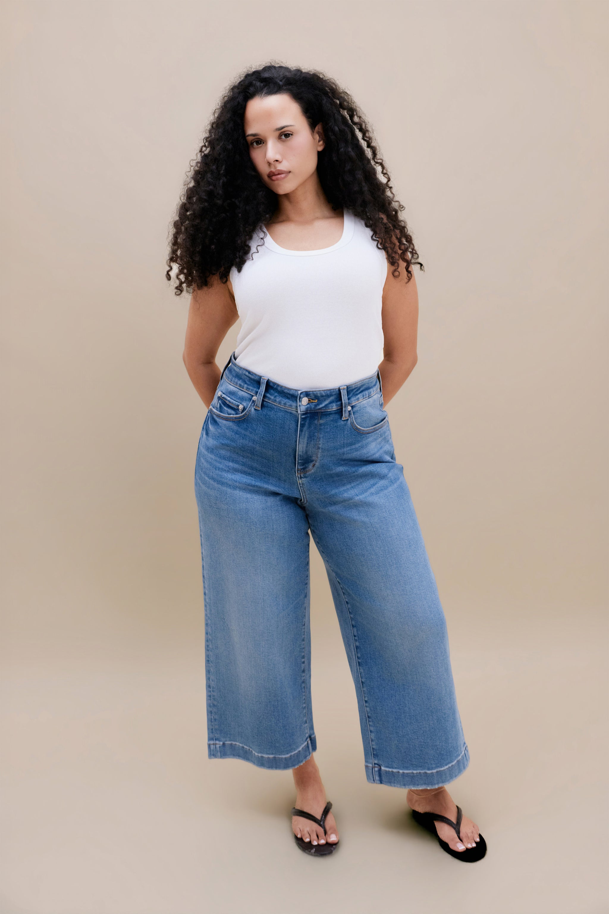 Woman wearing a white tank top with wide leg cropped jeans with a beige background | model-size: 16