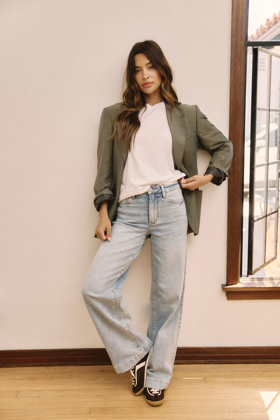 ADA Complaint: Shop Wide Leg Jeans.  Woman standing inside leaning against a white wall wearing a white tee, gray blazer, and light colored jeans.