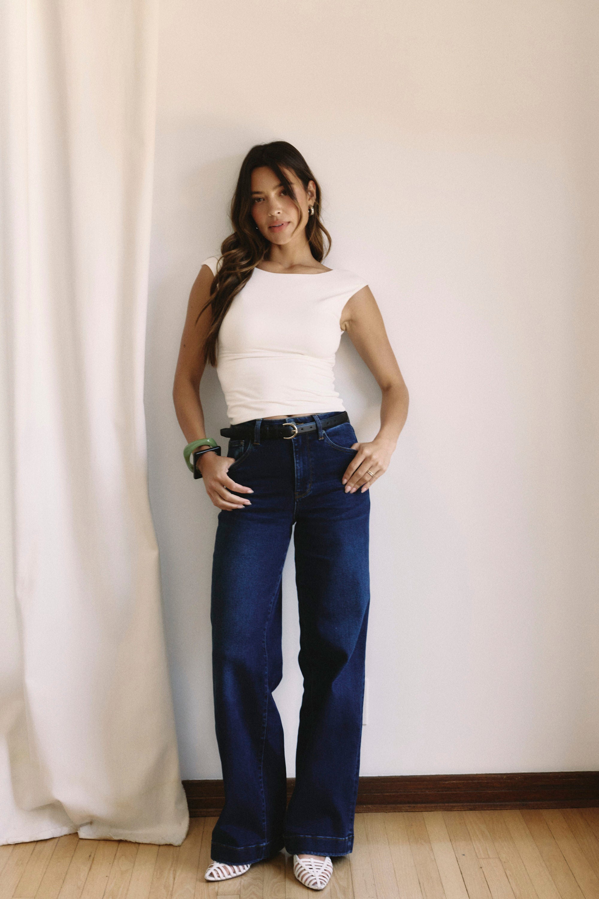 Woman wearing a white top and blue jeans standing against a white curtain  | model-size: 4