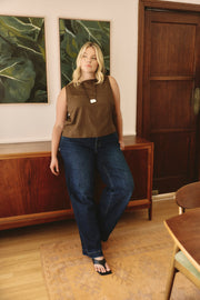 Woman in a brown sleeveless top and blue jeans standing in a room with wooden furniture and a large painting.