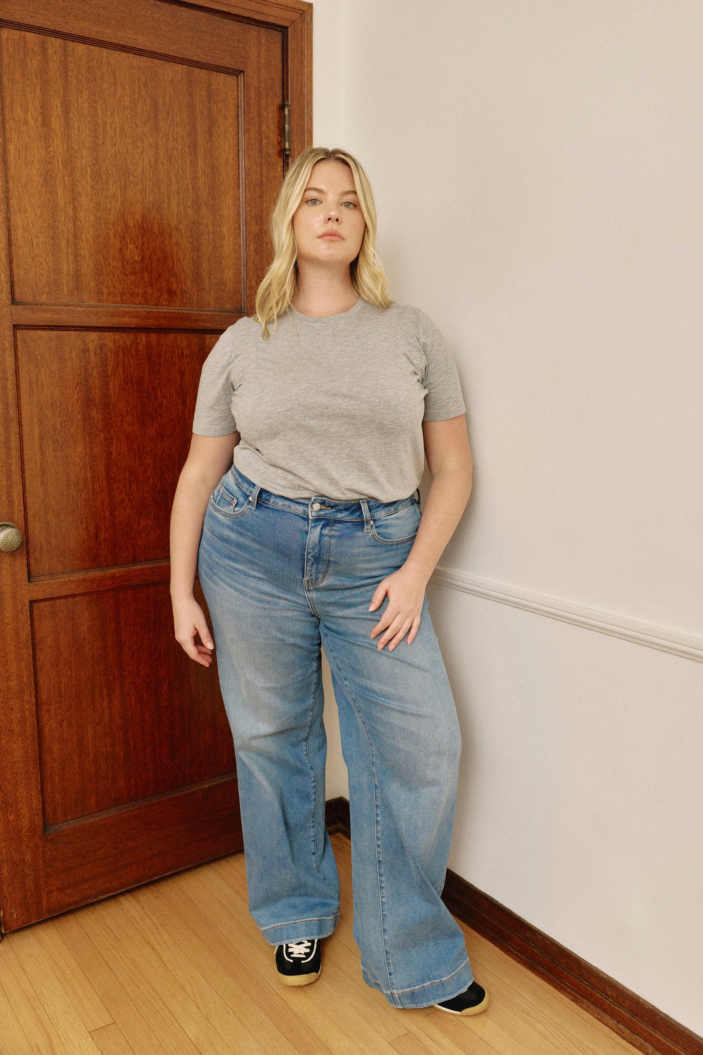 Person wearing a gray t-shirt and blue jeans standing in a room with wooden flooring and a wooden door.
