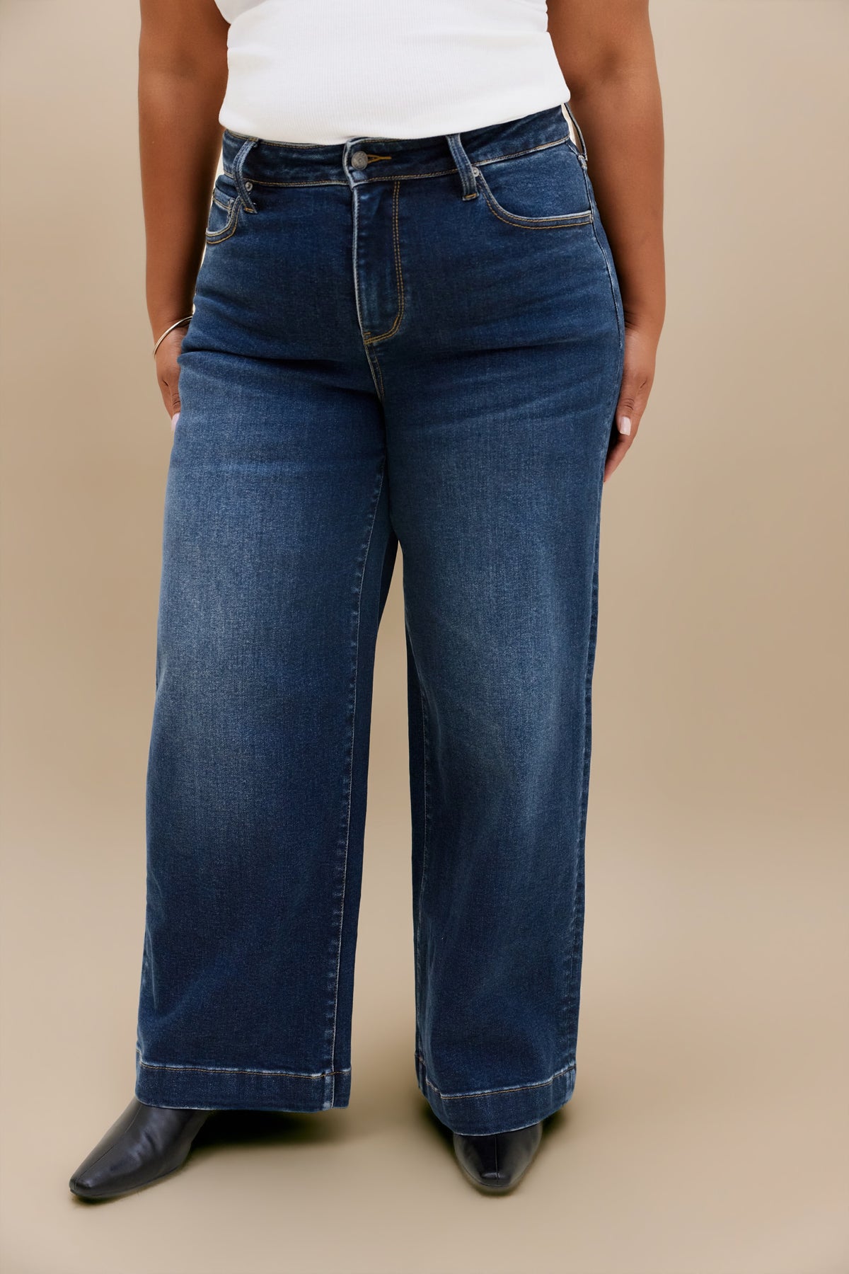 A person wearing dark blue wide-leg denim jeans with a white top and black pointed shoes, standing against a plain beige studio background.