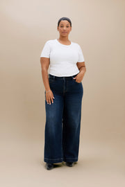 A woman modeling a pair of dark blue wide-leg jeans with a white short-sleeve t-shirt, standing against a solid beige studio background.