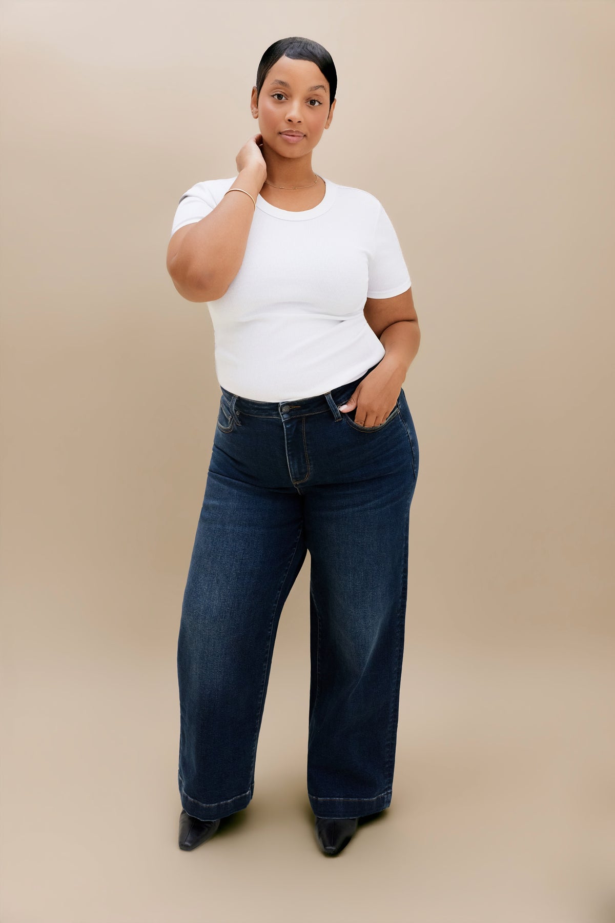 A woman modeling dark blue wide-leg jeans and a white t-shirt against a solid beige studio background.