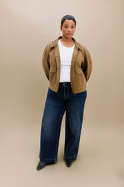A model stands against a solid beige studio background wearing a brown cropped jacket over a white top, dark blue wide-leg jeans, and black pointed shoes.