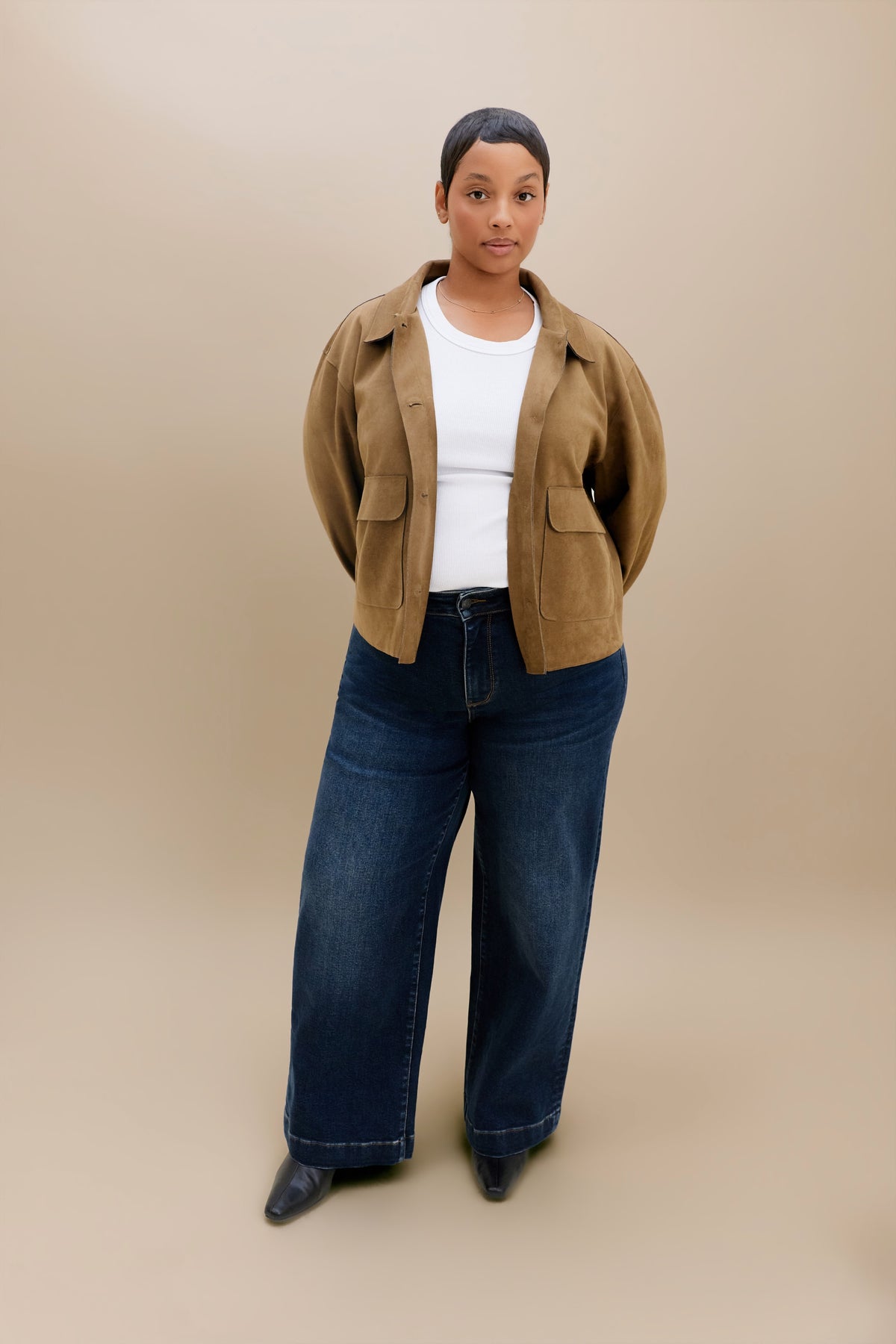 A model stands against a solid beige studio background wearing a brown cropped jacket over a white top, dark blue wide-leg jeans, and black pointed shoes.