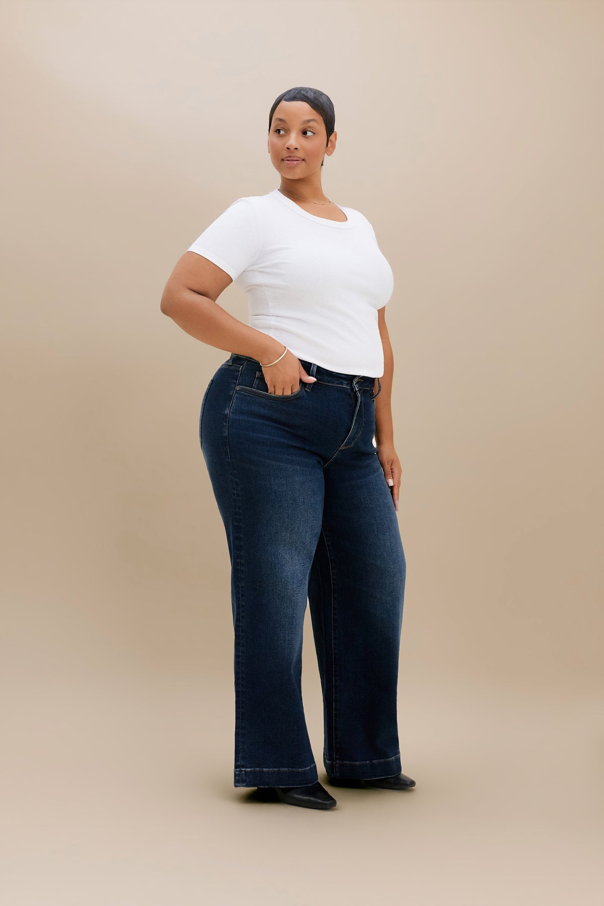A woman modeling dark blue wide-leg jeans with a white t-shirt, standing against a solid beige studio background.