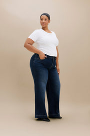A woman modeling dark blue wide-leg jeans with a white t-shirt, standing against a solid beige studio background.