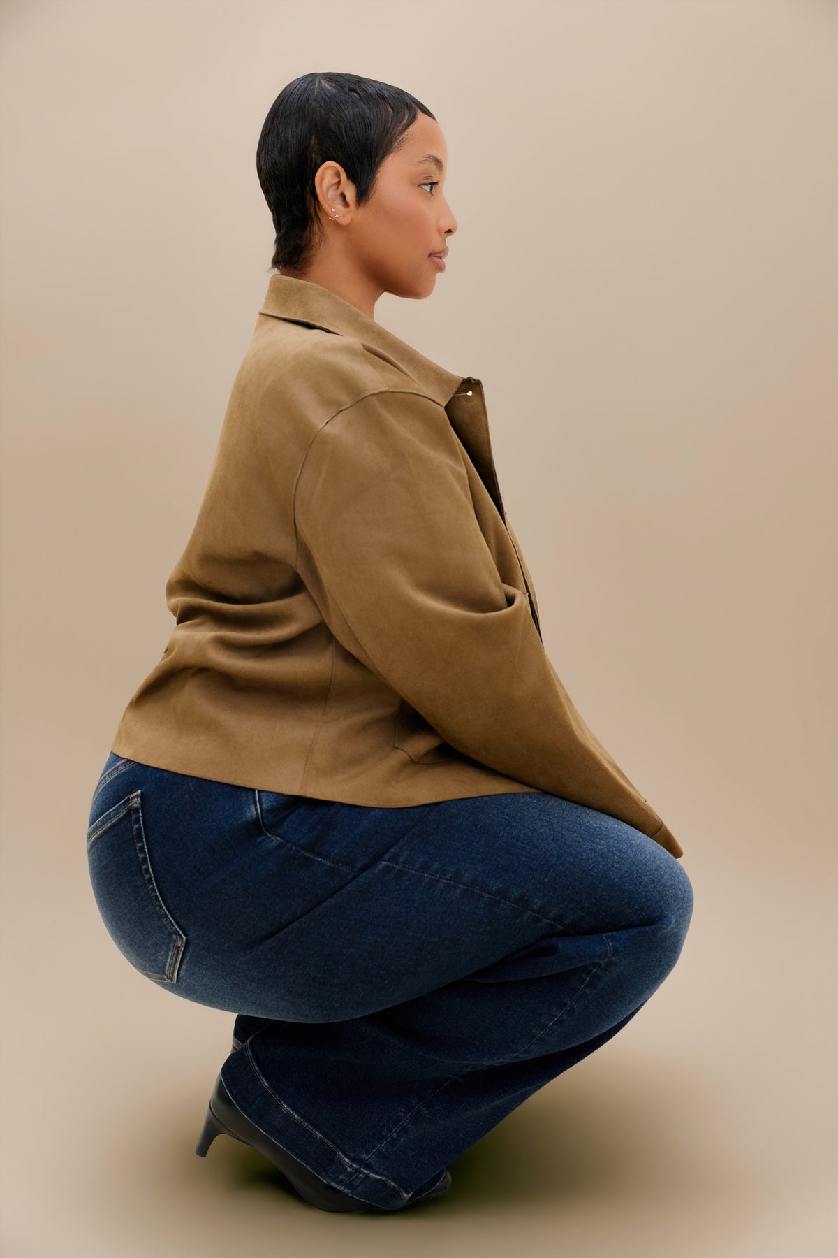 A model crouches in profile wearing a tan suede jacket and dark blue wide-leg jeans against a solid beige studio background.