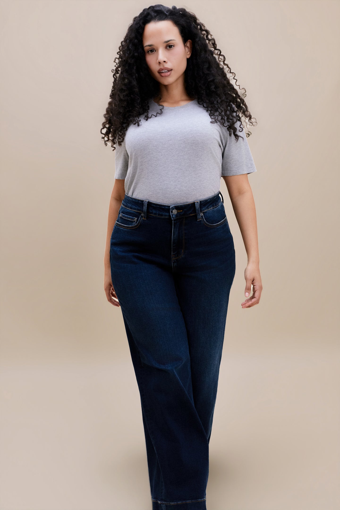Woman wearing a gray t-shirt and dark wide leg jeans standing with a beige background