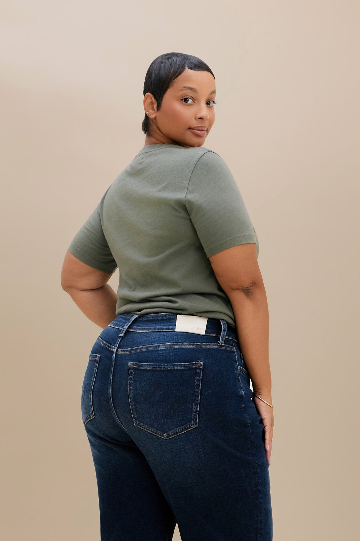 A woman wearing a muted sage green short-sleeve t-shirt and dark blue jeans, photographed from the back against a solid beige studio background.