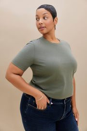 A woman modeling a short-sleeve crewneck t-shirt in sage green, paired with dark blue jeans, against a solid beige studio background.