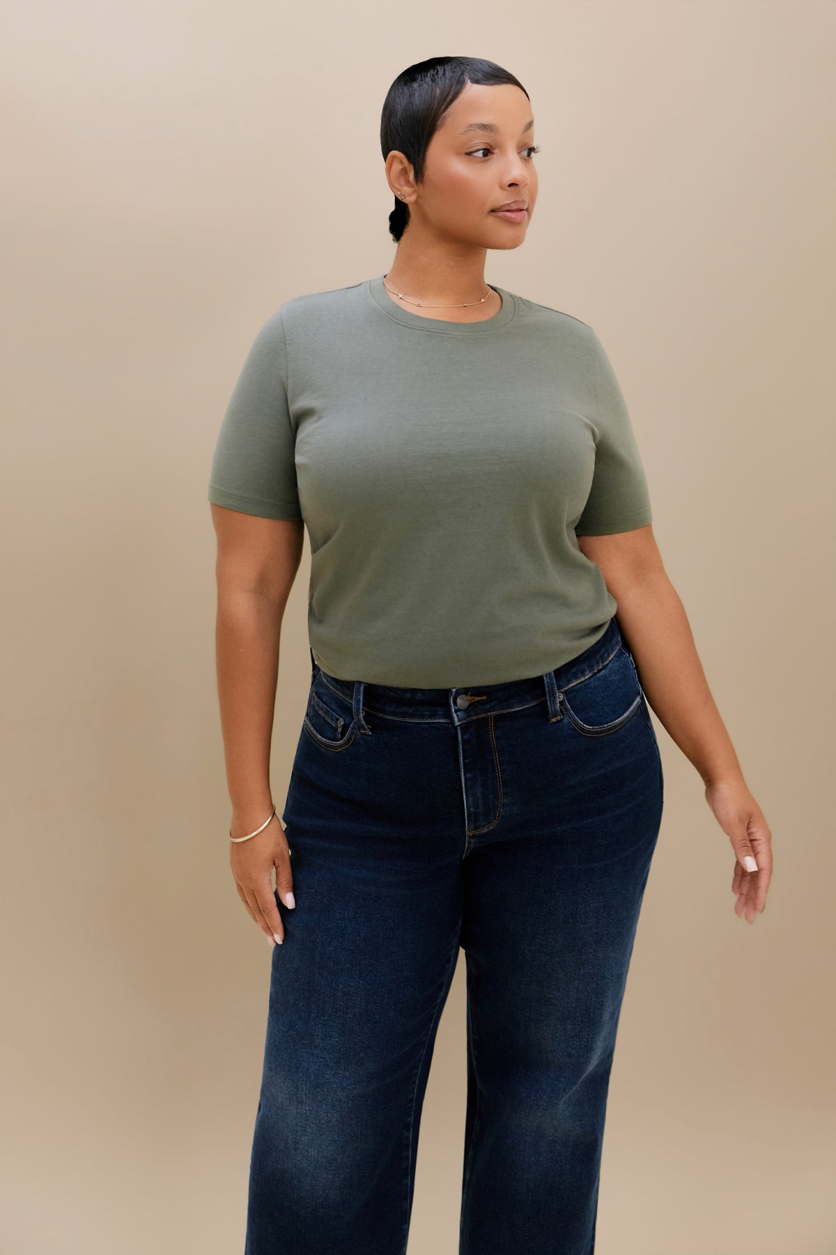 A woman modeling a short-sleeve crewneck t-shirt in sage green, paired with dark blue jeans, against a solid beige studio background.