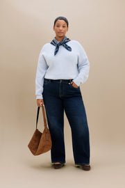 A woman stands against a plain beige studio background wearing a light blue knit sweater, dark striaght leg jeans, a navy patterned neck scarf, and dark flats, while holding a brown suede tote bag.
