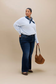 A woman modeling a light blue knit sweater, dark blue jeans, a navy patterned neck scarf, and brown shoes, while holding a brown suede shoulder bag against a solid beige studio background.