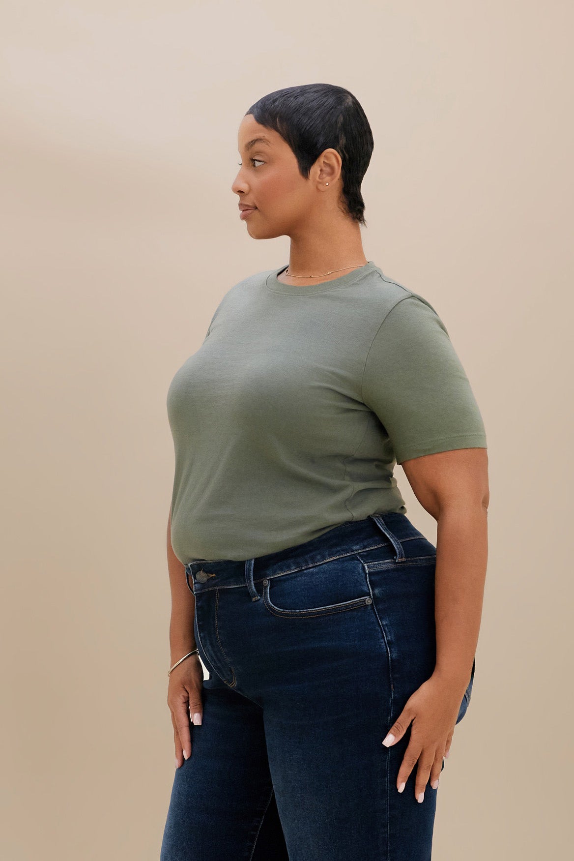A woman with short dark hair is shown in profile wearing a muted sage green short-sleeve crewneck t-shirt and dark blue jeans, standing against a solid beige studio background.