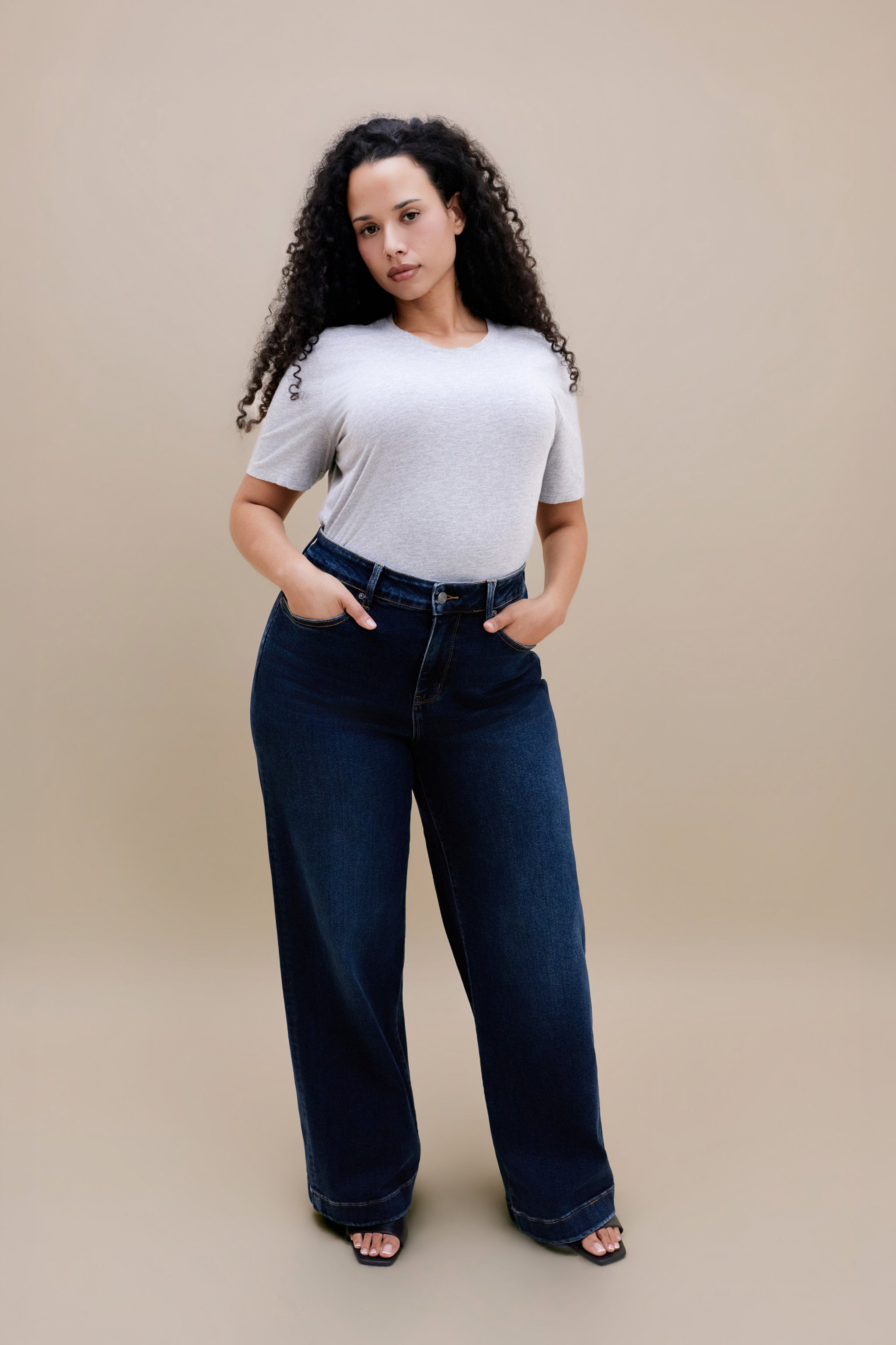 Woman wearing a gray t-shirt and dark wide leg jeans standing with a beige background | model-size: 16