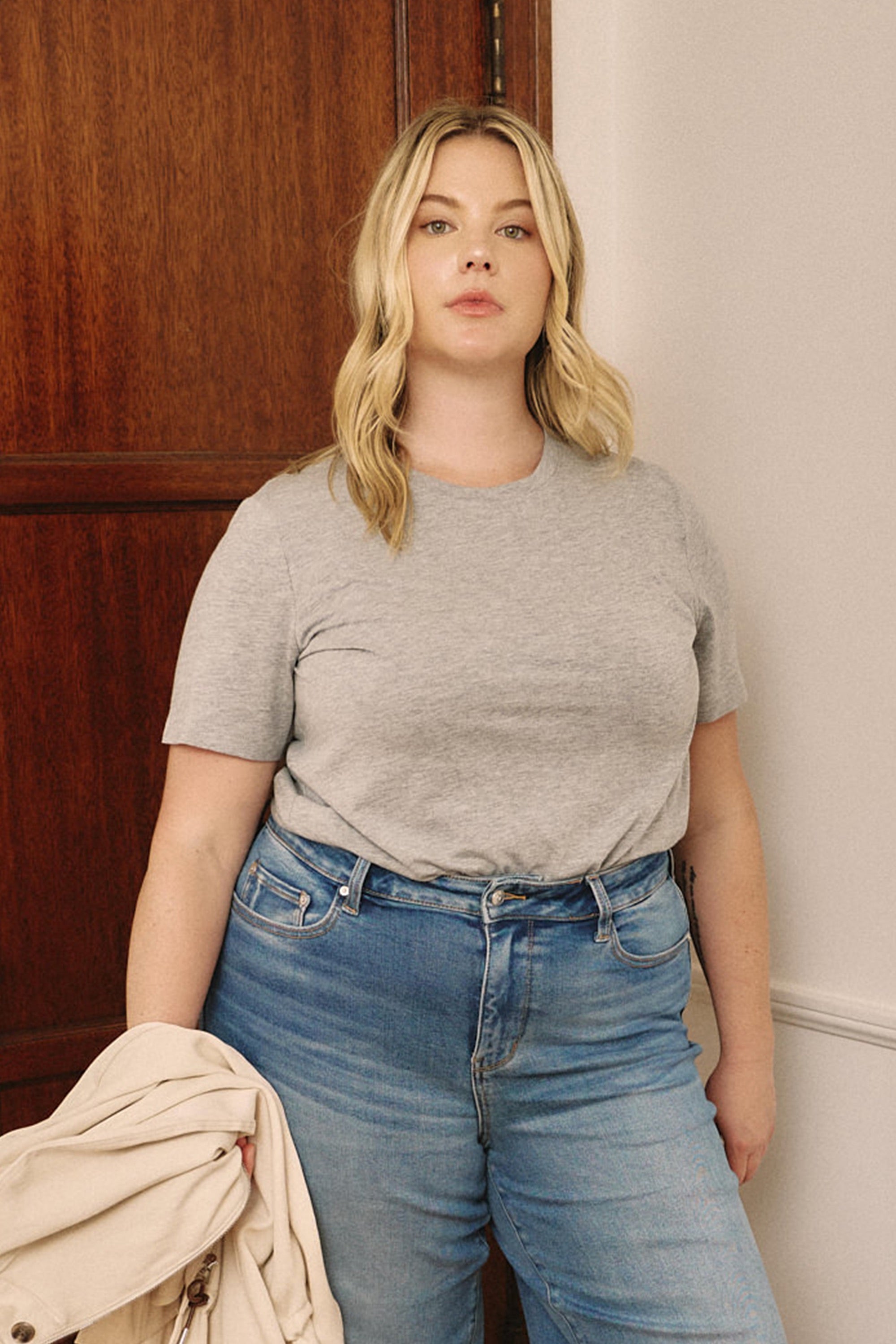 Woman wearing a gray t-shirt and blue jeans standing against a wooden door | model-size: 16