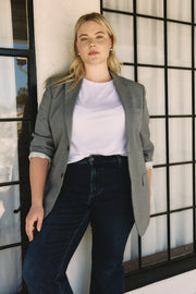 Woman wearing a gray blazer over a white shirt and dark jeans, standing against a windowed wall | model-size: 16