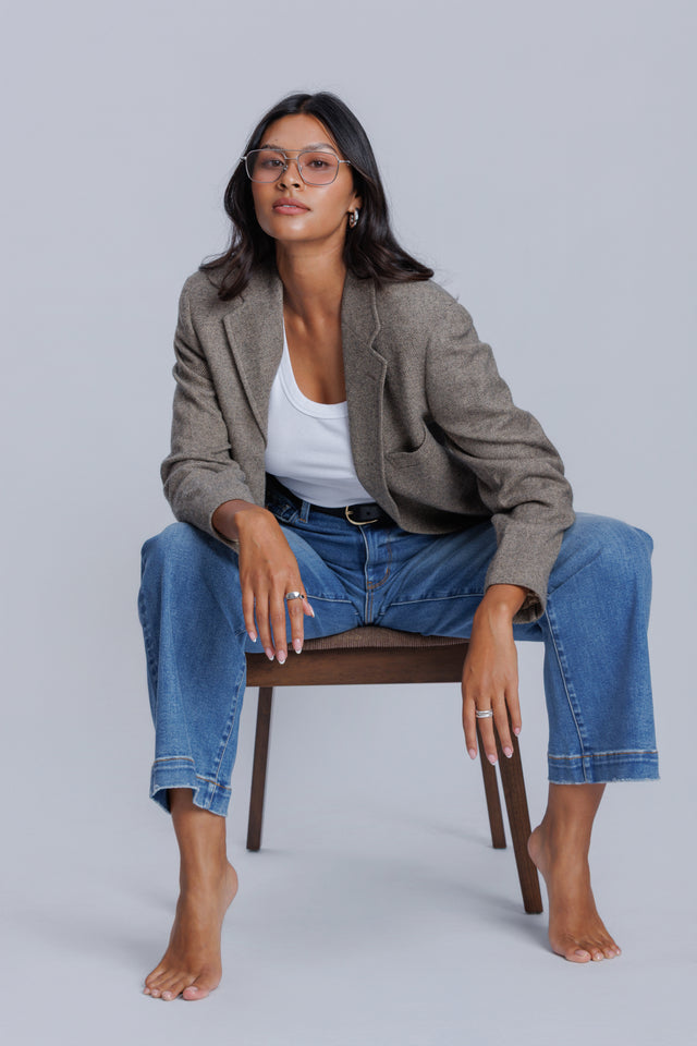 Woman sitting on a chair wearing a gray jacket, DePart white top, and blue jeans against a plain background