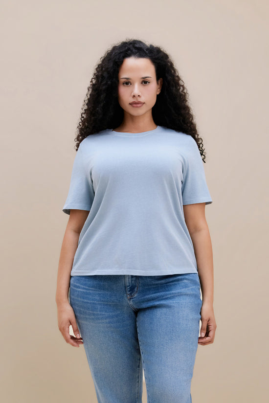 Woman wearing a blue crew neck tee and jeans with a beige background | model-size: 16