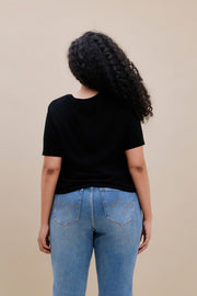 Back side of a woman wearing a black t-shirt and blue jeans against a beige background 