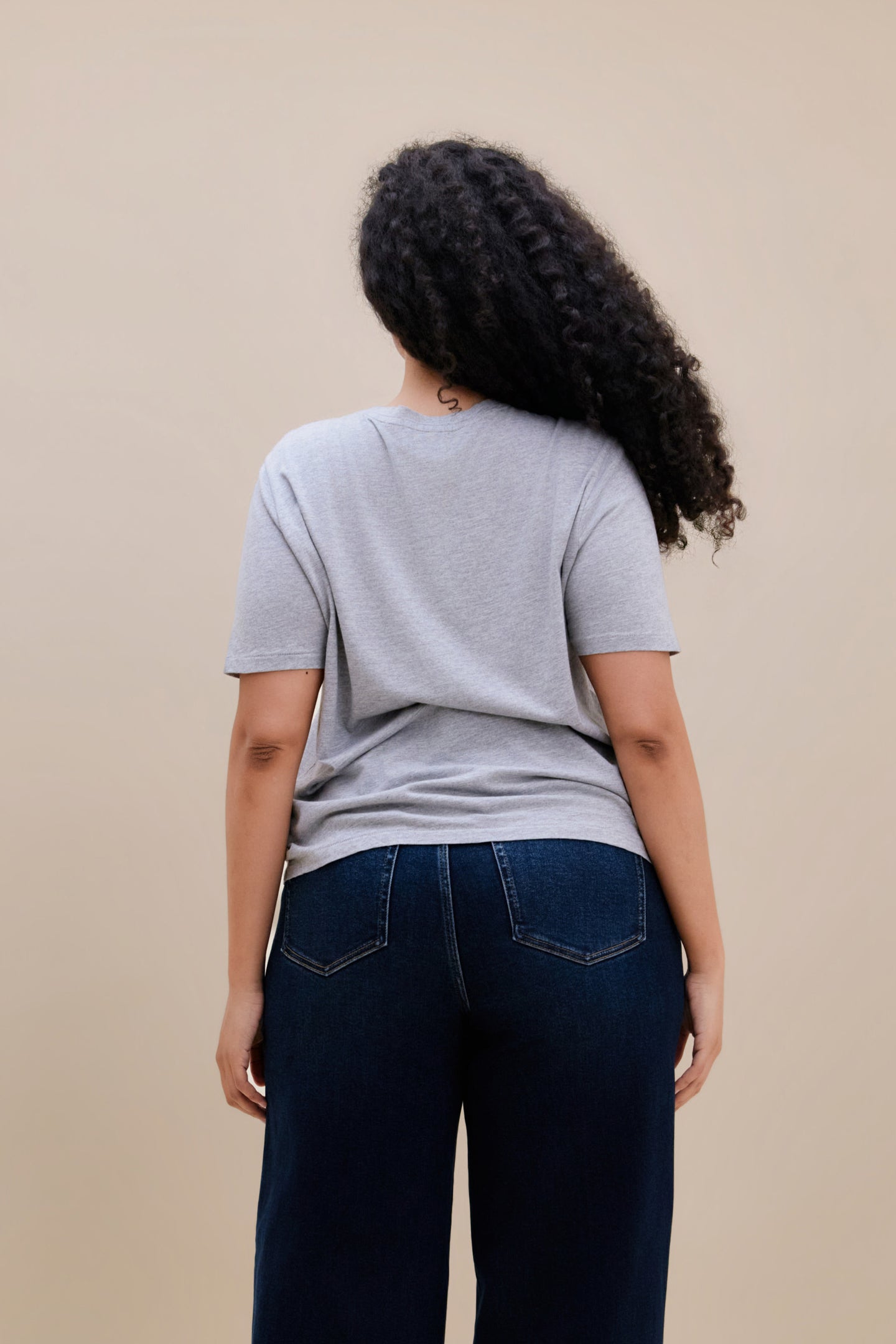 Backside of a woman wearing a light heather grey t-shirt  & dark jeans against a beige background
