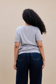 Backside of a woman wearing a light heather grey t-shirt  & dark jeans against a beige background