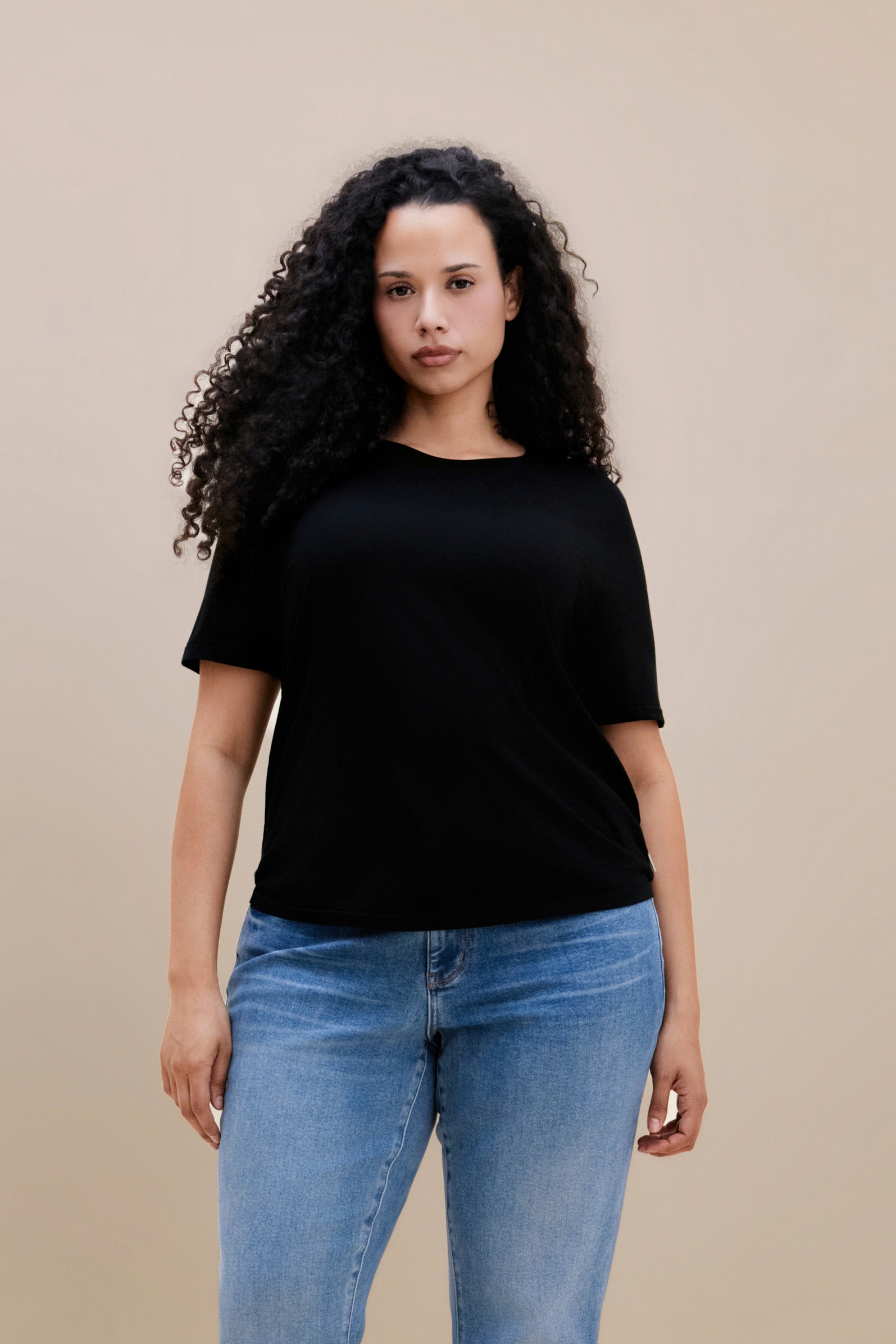 Woman wearing a black t-shirt with jeans against a beige background | model-size: 16
