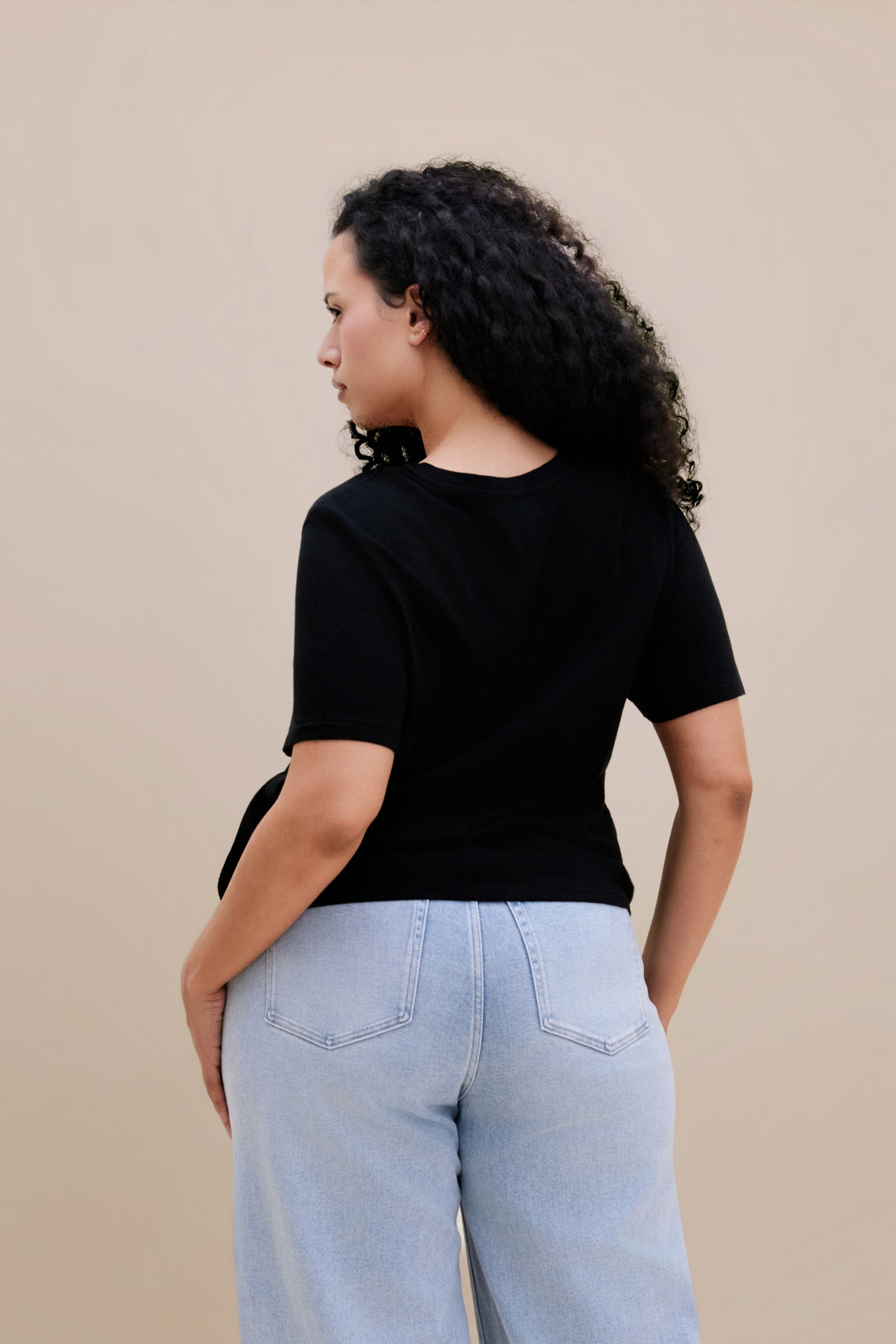 Backside of a woman wearing a black t-shirt and light blue jeans against a beige background 