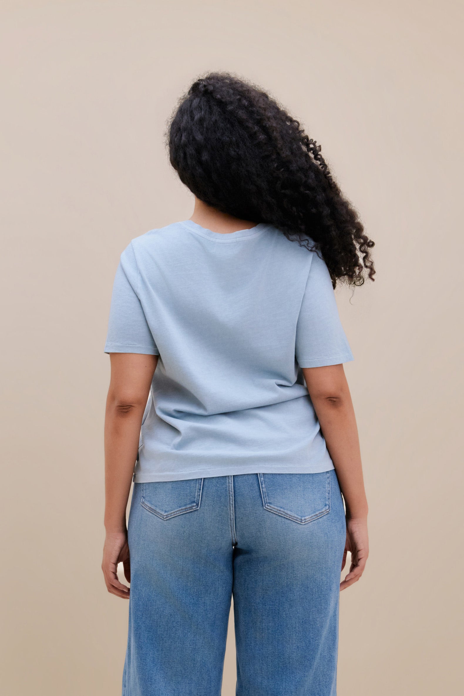 Backside of a woman wearing a blue t-shirt and jeans with a beige background